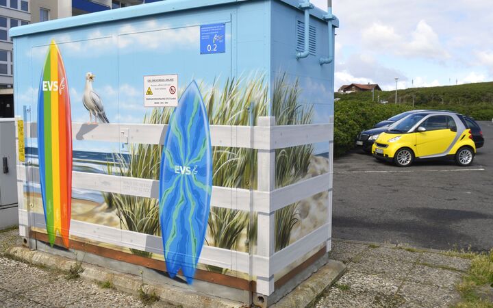 Eine bemalte Trafostation mit Surfbrettern, die an einem Zaun lehnen